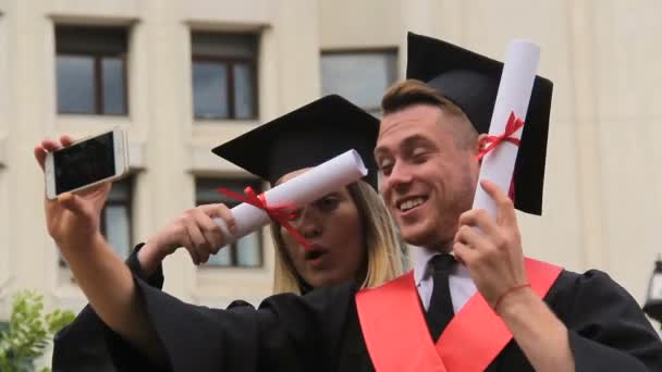 Feminino e masculino graduados em vestidos acadêmicos e bonés de filmagem de vídeo no telefone — Vídeo de Stock