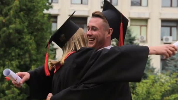 Mejores amigos en vestidos académicos y sombreros abrazando y sonriendo en el día de la graduación — Vídeos de Stock