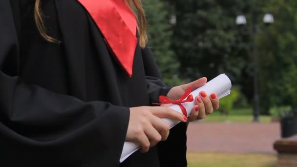 Pleased female graduate in academic dress holding diploma, bright future