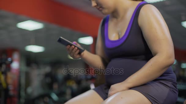 Товста нездорова жінка з великим животом перевіряє додаток для смартфона в спортзалі, втрата ваги — стокове відео