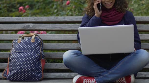 Mujer birracial sonriente sentada en el banco, sosteniendo el portátil y hablando por teléfono celular — Vídeos de Stock