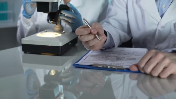 Lab worker filling paper forms, his assistant viewing samples under microscope — Stock Video