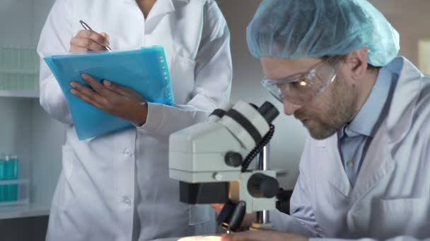 Biólogo estudiando muestras bajo microscopio, asistente anotando comentarios — Vídeos de Stock