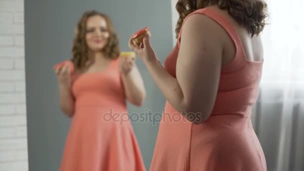 Happy and satisfied with oversize body girl eating donuts in front of mirror — Stock Video