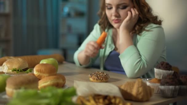 Грустная толстая девушка ест морковь, смотрит на пончики и фаст-фуд, диеты — стоковое видео
