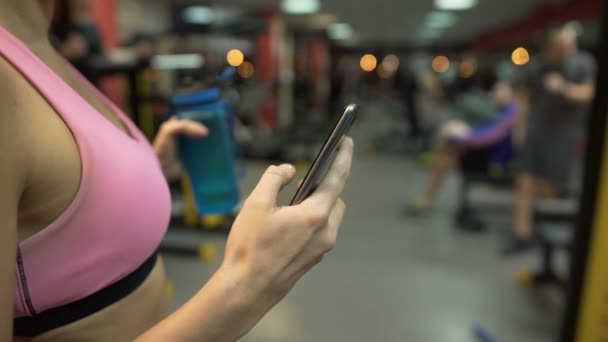 Chica teniendo un descanso del ejercicio en el gimnasio, comprobando el teléfono celular, botella de agua en la mano — Vídeos de Stock