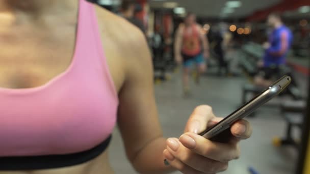 Mujer joven desplazando la pantalla del teléfono inteligente en el gimnasio, la creación de la aplicación de entrenamiento — Vídeos de Stock