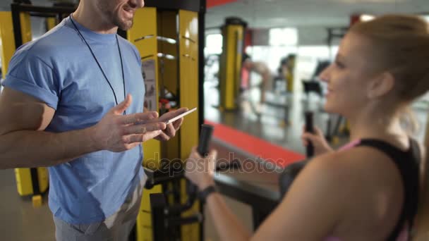 Trener, omawiając plan treningowy z jego kobieta klienta, para flirtuje w siłowni — Wideo stockowe