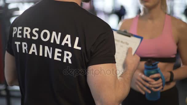 Entrenador de fitness explicando programa de entrenamiento y horario a cliente femenina en gimnasio — Vídeos de Stock