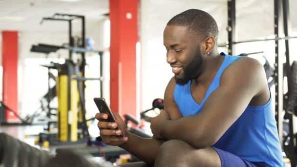 Feliz homem afro-americano sentado no ginásio e assistindo vídeo no celular — Vídeo de Stock