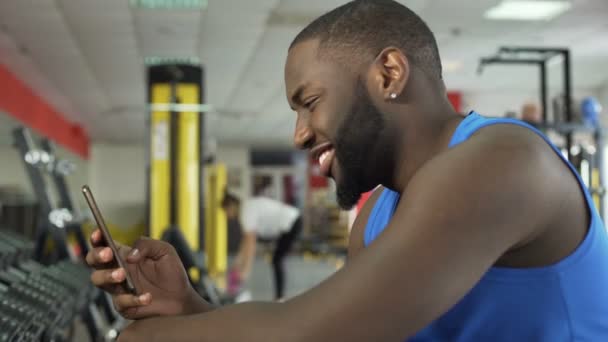 Atleta sonriente con éxito desplazando fotos en el teléfono inteligente en el club de fitness — Vídeo de stock