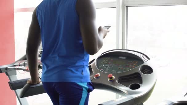 Afro-American mannen gå på löpband och kontrollera sin smartphone i gymmet — Stockvideo