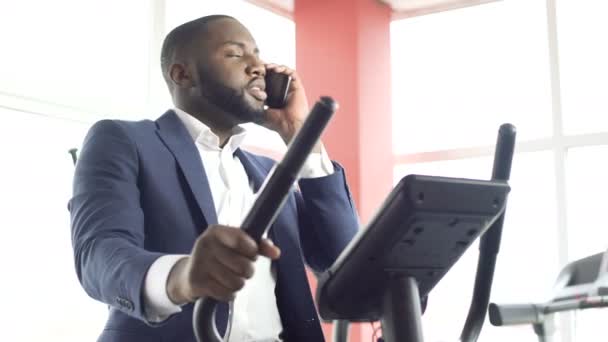 Serious male in suit exercising on stationary bike and talking on cellphone — Stock Video