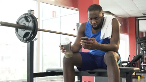 Мужчина сидит в спортзале, пьет белковый коктейль и прокручивает по телефону — стоковое видео