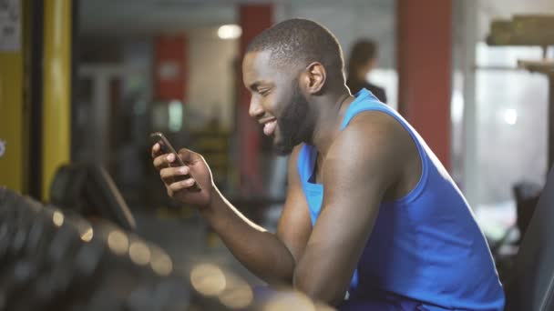 Hombre joven escribiendo mensaje en el teléfono inteligente con sonrisa en la cara, descanso durante el entrenamiento — Vídeo de stock