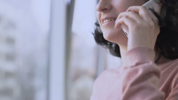 Γυναίκα, που χαμογελά ενώ έχοντας συνομιλία μέσω του κινητού τηλεφώνου, παράθυρο στο υπόβαθρο — Αρχείο Βίντεο