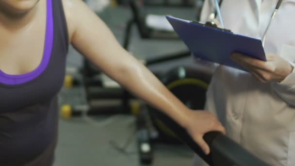 Спітніла товста жінка тренується на біговій доріжці, лікар бере результати, реабілітація — стокове відео