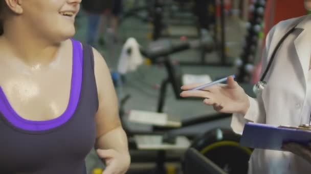 Chica obesa haciendo ejercicio en la cinta de correr en el gimnasio, hablando con la enfermera a un lado, reloj de peso — Vídeo de stock