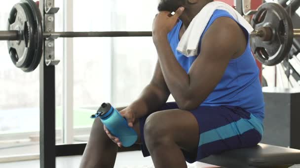 Joven hombre sentado en el gimnasio, sosteniendo la botella de agua, limpiándose la cara con una toalla — Vídeo de stock