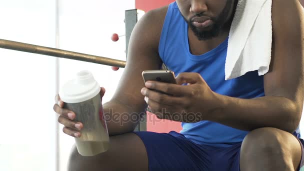 Masculino sentado no ginásio, segurando coquetel de proteína e telefone celular, feed de notícias sociais — Vídeo de Stock