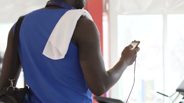 Афроамериканец, стоящий в спортзале, прокручивающий экран мобильного телефона, финиш — стоковое видео