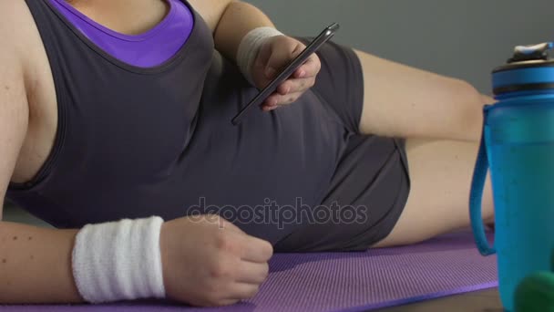 Giovane donna sovrappeso sdraiata su tappetino yoga, schermo del cellulare a scorrimento, pigrizia — Video Stock