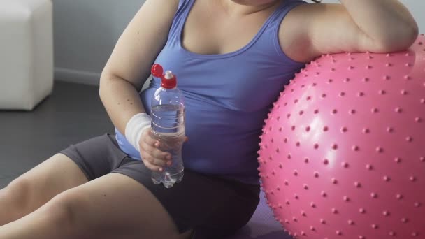 Gordura jovem sentada no chão bebendo água, inclinando-se na bola de fitness, treino — Vídeo de Stock