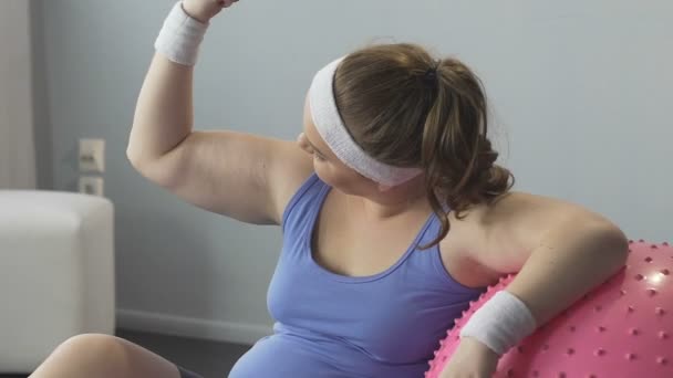 Fille en surpoids assis sur le sol appuyé sur la balle de remise en forme, flexion des bras satisfaits — Video