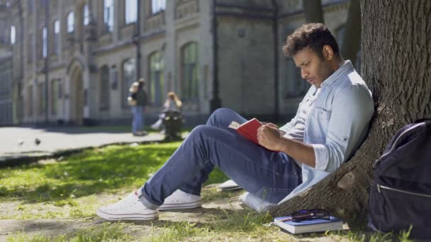 Multiracial ung kille sitter under träd, Läsning intressant bok, bookworm — Stockvideo