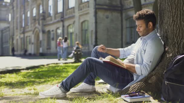 Multinacional chico sentado bajo el árbol, libro de lectura mirando a su alrededor, tiempo libre — Vídeos de Stock