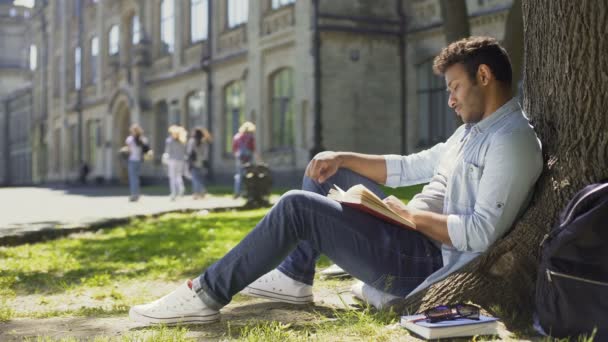 Jeune homme assis sous un arbre avec un livre regardant autour, ayant des pensées agréables — Video