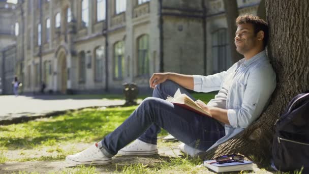 Mixed-race guy sitting under tree, waving and smiling at somebody, friendly — Stock Video