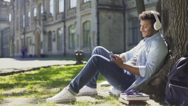 Homme dans un casque assis sur l'herbe et appuyé contre l'arbre, écoutant de la musique — Video