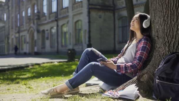 Kvinnlig student sitter på gräset, lutad mot trädet, lyssna på musik, koppla av — Stockvideo