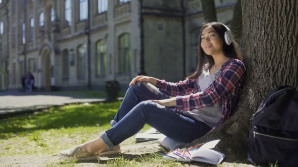 Mujer joven multinacional en auriculares sentados bajo el árbol, escuchando música — Vídeos de Stock