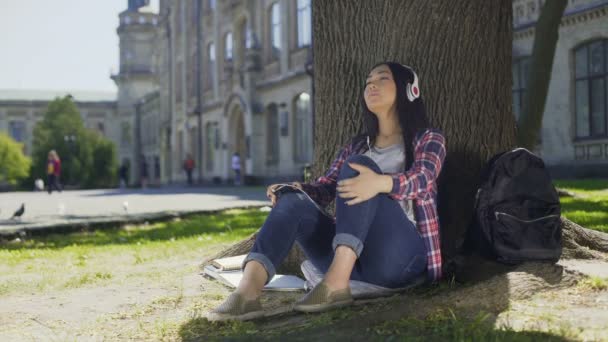 Asiática hembra en auriculares disfrutando de la música, sentado bajo el árbol, banda favorita — Vídeos de Stock