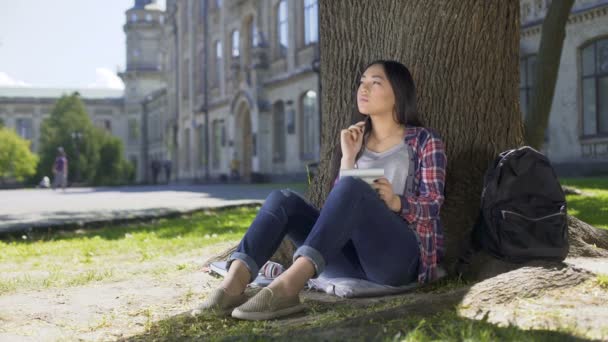 Estudiante de intercambio sentado bajo el árbol, sosteniendo el cuaderno, pensando, lista de check-in — Vídeos de Stock