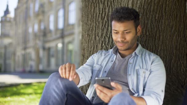 Mixed young man sitting under tree, scrolling down cellphone screen, laughing — Stock Video