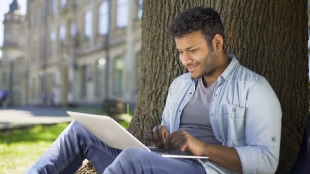 Πολυεθνική νεαρός άνδρας κάθεται κάτω από το δέντρο, πληκτρολογώντας σε φορητό υπολογιστή, χαμογελώντας, κουβεντιάζοντας — Αρχείο Βίντεο
