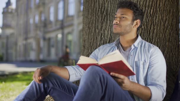 Молодой человек сидит под деревом чтение книги и мышление, литература, фантастика — стоковое видео