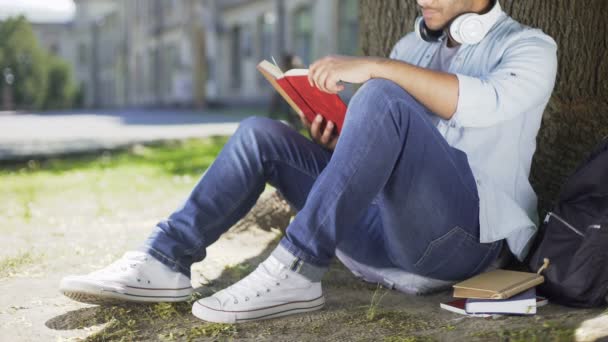 Wielonarodowych facet siedzący pod drzewem, czytanie książki, książki kochanka, czas wolny — Wideo stockowe