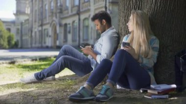 Çok uluslu adam ve kız telefonları ile ağacın altında oturan kısa göz değişimi