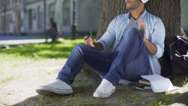 Πολυφυλετικές αρσενικό ακουστικά που κάθεται κάτω από το δέντρο, να ακούτε την αγαπημένη μουσική — Αρχείο Βίντεο