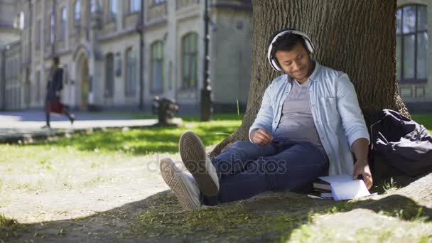 Tipo de raza mixta sentado bajo el árbol en los auriculares escribiendo en el cuaderno, escritor de canciones — Vídeo de stock