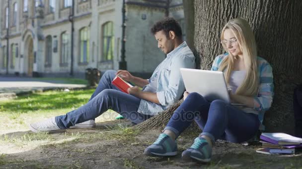 Joven sentado bajo el árbol con libro, mirando a la chica con el ordenador portátil, indeciso — Vídeos de Stock