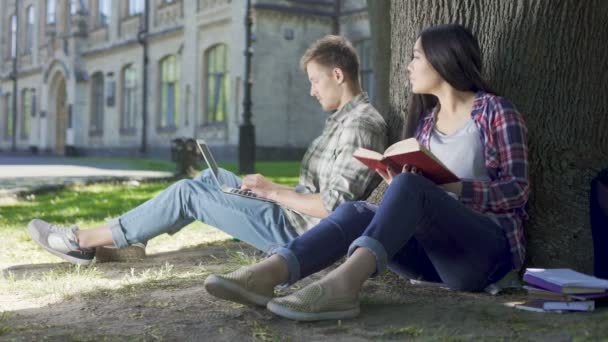 Asiatisk tjej läser bok under träd och tittar på killen med laptop, blyghet — Stockvideo