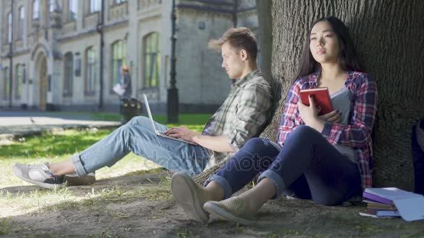 Multinationale meisje zittend onder de boom, holding boek, kijken naar vent, gevoelens — Stockvideo