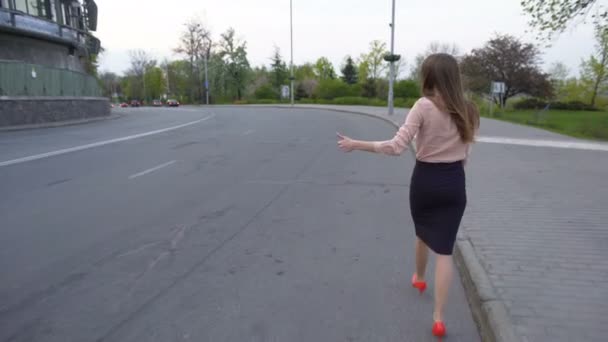 Nuori kaunis liiketoiminnan nainen kävelee tiellä ja yrittää liftata auton — kuvapankkivideo
