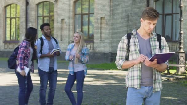 Einsamer männlicher Student schaut glücklich lachende Mitschüler an, die in der Nähe des College stehen — Stockvideo