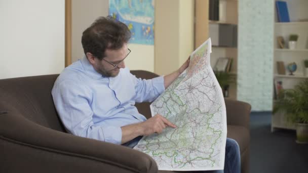 Glada mannen sitter på soffan och att välja resort city på karta, äventyrare — Stockvideo
